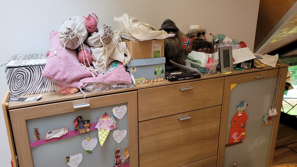 Ein überfüllter Holzschrank, bedeckt mit Taschen, Kisten, Spielzeug und Papier. Kindliche Zeichnungen schmücken den Schrank und erzeugen eine verspielte, chaotische Szene.