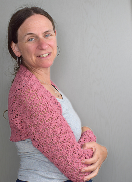 Sanft lächelnde Frau mit einer rosa gehäkelten Stola über einem grauen Oberteil vor einem schlichten Hintergrund. Ihre entspannte Haltung vermittelt eine warme, freundliche Ausstrahlung.