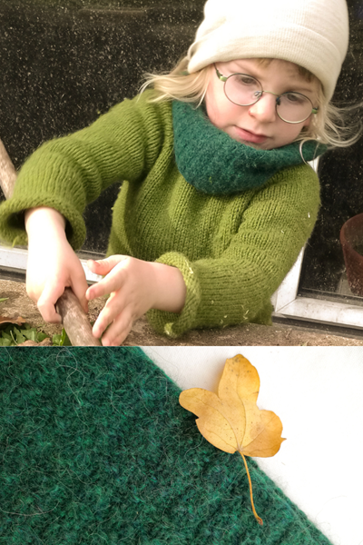 Ein Kind mit Brille und weißer Mütze trägt einen grünen Strickpullover und einen grünen Loop- Schal und beugt sich neugierig nach vorne. Darunter liegt ein gelbes Blatt auf dem grünen Loop.