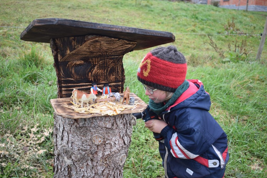 Ein Kind in Winterjacke und Mütze betrachtet aufmerksam eine kleine hölzerne Krippenszene auf einem Baumstumpf. Die Umgebung ist eine grasbewachsene Außenfläche.