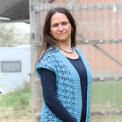 Eine Frau mit langen braunen Haaren steht im Freien, trägt eine blaue gehäkelte Weste und ein schwarzes Shirt. Sie lächelt sanft, vor einem Gewächshaus und einem Camper. Ruhige, ländliche Umgebung.