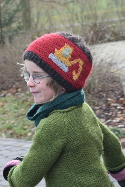 Ein Kind mit Brille und einer bunten Häkelmütze mit Bagger-Motiv, gekleidet in einen grünen Pullover und einen Schal, lächelt fröhlich, während es draußen über graue Pflastersteine läuft.