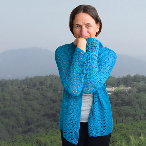 Eine Frau steht vor einer üppigen, grünen Berglandschaft und trägt eine leuchtend blaue gehäkelte Strickjacke. Sie lächelt sanft und vermittelt eine ruhige, fröhliche Stimmung.