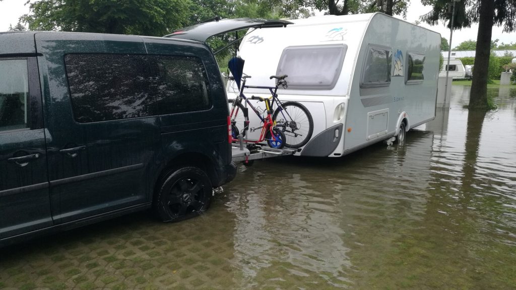 Ein Van mit angehängtem Wohnwagen und befestigtem Fahrrad steht auf einer überfluteten Straße. Die Stimmung ist angespannt, der Himmel ist bewölkt und Wasser umgibt die Fahrzeuge.