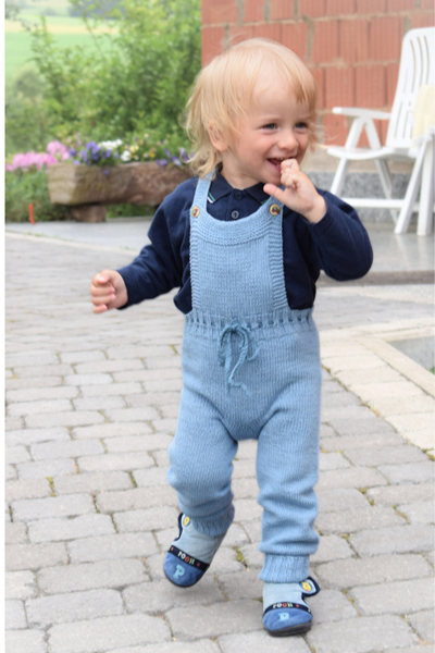 Ein fröhliches Kleinkind mit blonden Haaren läuft über einen Kopfsteinpflasterweg, gekleidet in blaue Latzhose und marineblaue Schuhen. Im Hintergrund sind grünes Gras und bunte Blumen zu sehen.