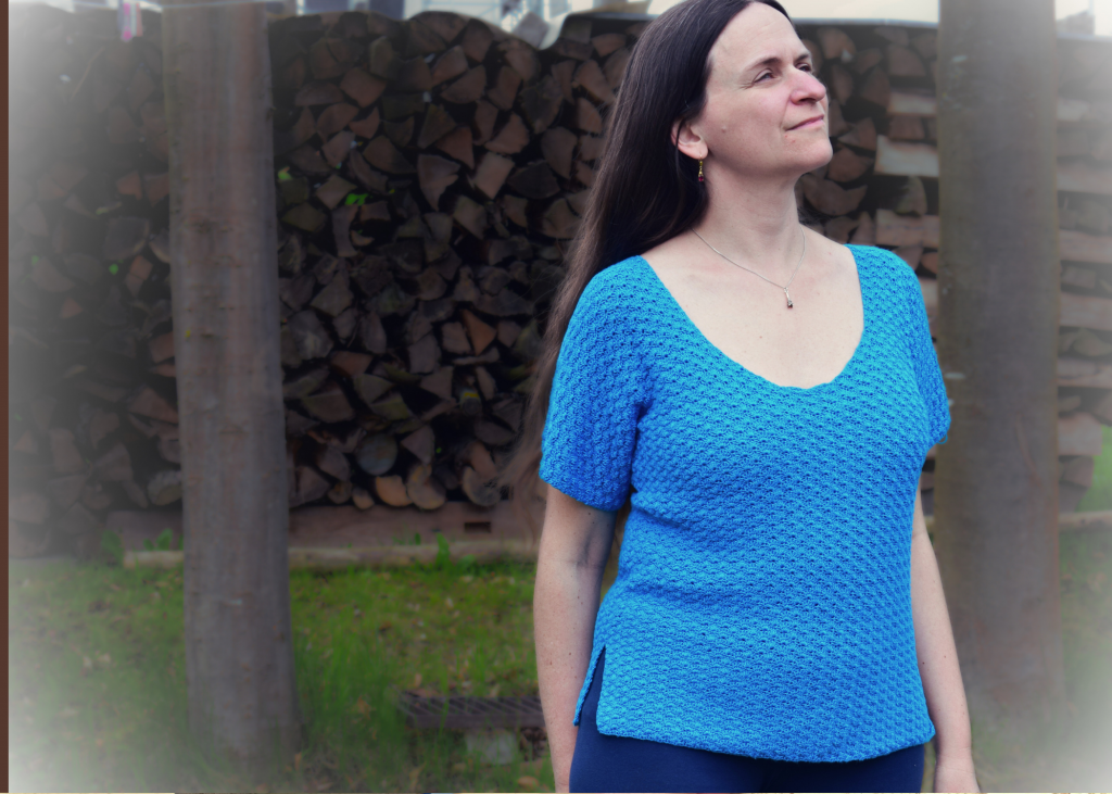 Eine Frau in einem blauen Häkeloberteil steht im Freien und blickt mit einem gelassenen Ausdruck nach oben. Im Hintergrund sind ein Holzstapel und Bäume zu sehen, die eine ruhige, natürliche Atmosphäre vermitteln.