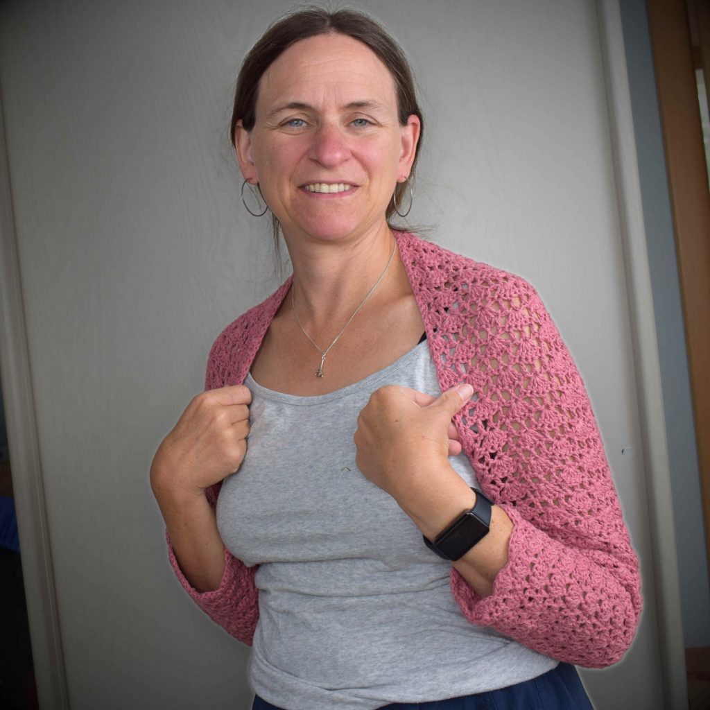 Lächelnde Frau mit einem rosa gehäkelten Bolero über einem grauen Shirt. Sie hält den Bolero an den Schultern fest. Der Hintergrund ist ein neutraler Innenraum.