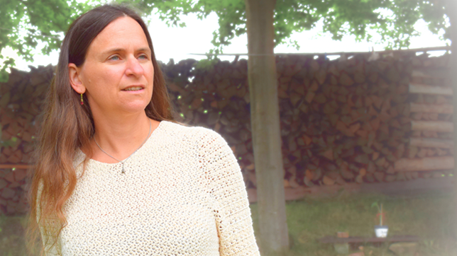 Eine Frau mit langen Haaren steht im Freien vor gestapeltem Brennholz. Sie trägt einen hellen Häkelpullover und blickt nachdenklich nach rechts. Im Hintergrund sind Bäume zu sehen.