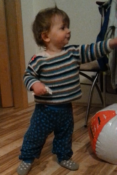 Ein Kleinkind in einem gestreiften Pullover und blauen Hosen zeigt neugierig in einem gemütlichen Raum mit Holzboden, neben dem eine große Tasche mit Kürbis-Motiv steht.