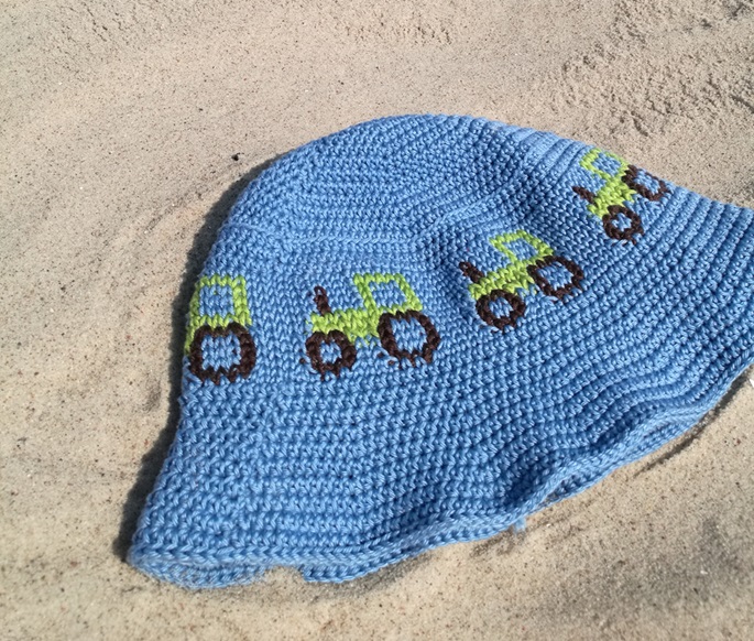 Eine blaue, gehäkelte Mütze mit grünen und braunen Traktor-Motiven liegt auf sandigem Boden und vermittelt ein verspieltes, sommerliches Gefühl