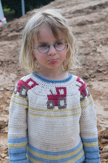 Ein Kind mit Brille und Schmollmund steht auf einem Feldweg und trägt einen gehäkelten Pullover mit roten Traktor-Motiven. Die Szene wirkt verspielt, aber leicht trotzig.