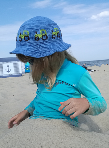 Ein Kind mit einem blauen gehäkelten Sonnenhut mit Traktormuster und einem türkisfarbenen Shirt sitzt am Sandstrand. Im Hintergrund sind eine Strandhütte und das Meer zu sehen.